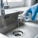 Image of a gloved hand pouring liquid from a bottle labeled acetone directly into a stainless steel kitchen sink drain, visualizing the common question regarding safe home disposal and the potential risks to plumbing and the environment.