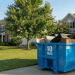 Blue 10 cubic yard dumpster sitting in a residential driveway filled with household junk and furniture, illustrating the actual size dimensions and capacity for a Jiffy Junk rental.