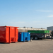 A wide shot of the Jiffy Junk Dumpster & Trailer Rentals yard under a clear blue sky, featuring a lineup of different sized roll-off dumpsters (red 40yd, blue 20yd, green 10yd) and two utility trailers on a gravel surface. The prominent sign and varied equipment illustrate Jiffy Junk as the best cheap local big and small roll off dumpster and trailer rentals near me, showcasing that Jiffy Junk has the right size at the right price for any project.