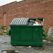 View of a brand new green 2 yard dumpster placed on an asphalt surface in front of a brick wall.
