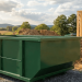 A realistic wide-angle shot of a clean, dark green 5-yard roll-off dumpster sitting on a gravel surface at a residential construction site.