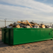 view of a large green 8 yard dumpster rental filled with construction debris and cardboard, sitting on a gravel lot near a chain-link fence under a blue sky.