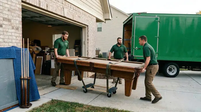 A view of a three professional carefully moving a large, ornate, wood-framed pool table on a wheeled moving dolly.