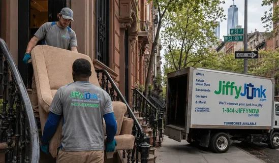 A Jiffy Junk crews carefully navigates a New York City brownstone stairwell while removing old furniture, with a branded Jiffy Junk truck visible at the curb, illustrating the professional junk hauling and pickup services that NYC residents trust across Manhattan, Brooklyn, and Queens.