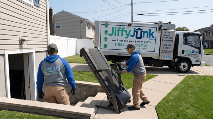 Two professional Jiffy Junk team members wearing branded Jiffy Blue and Jiffy Teal uniforms carefully maneuver a heavy folded treadmill out of a basement doorway toward a well-maintained, branded truck, illustrating the professional approach to bulky exercise equipment removal.